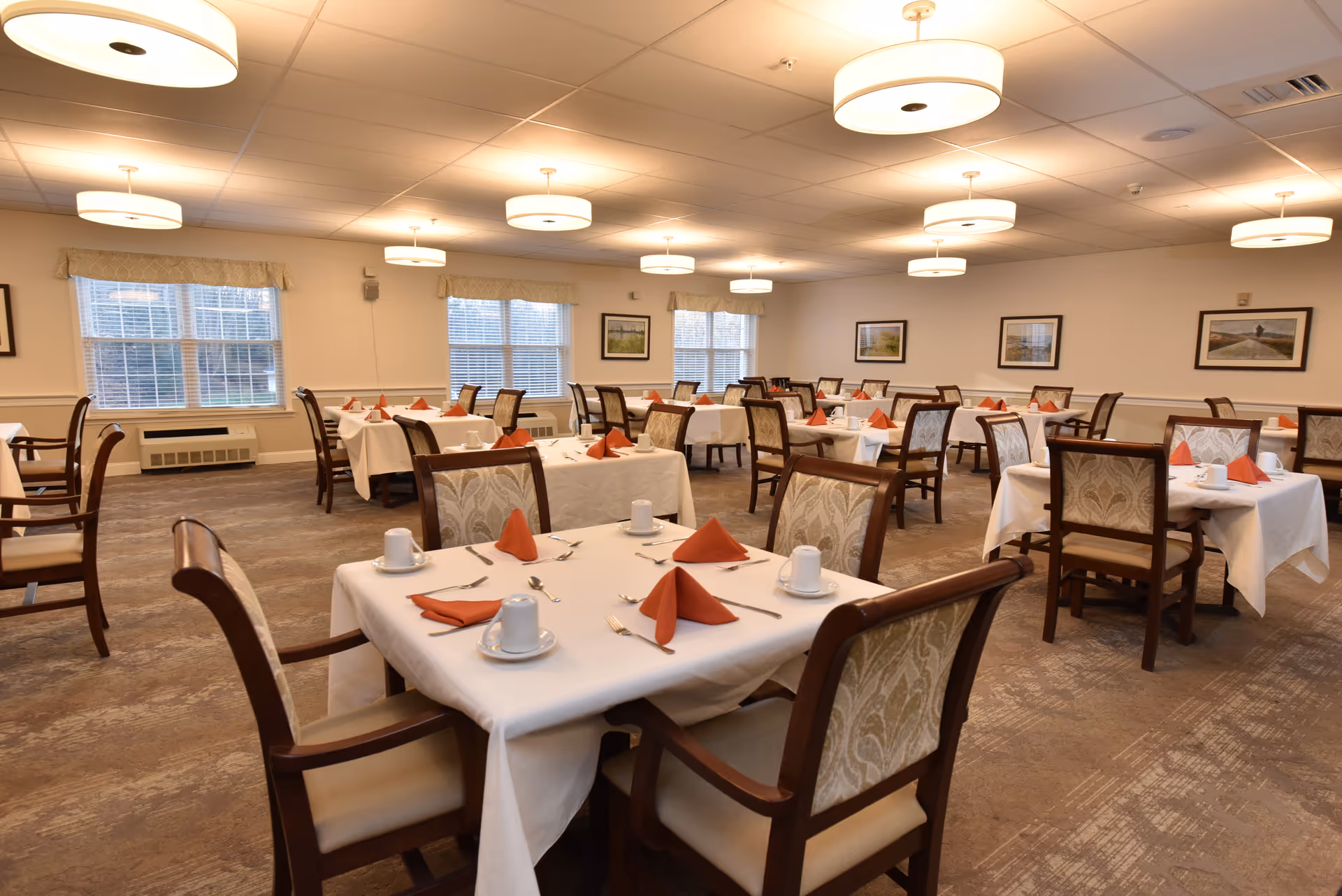 A spacious dining room with multiple tables covered in white tablecloths, each set with white cups, saucers, silverware, and orange folded napkins. The room has several large windows with beige valances, framed pictures on the walls, and ceiling lights providing warm illumination. The chairs have wooden frames with patterned upholstery.