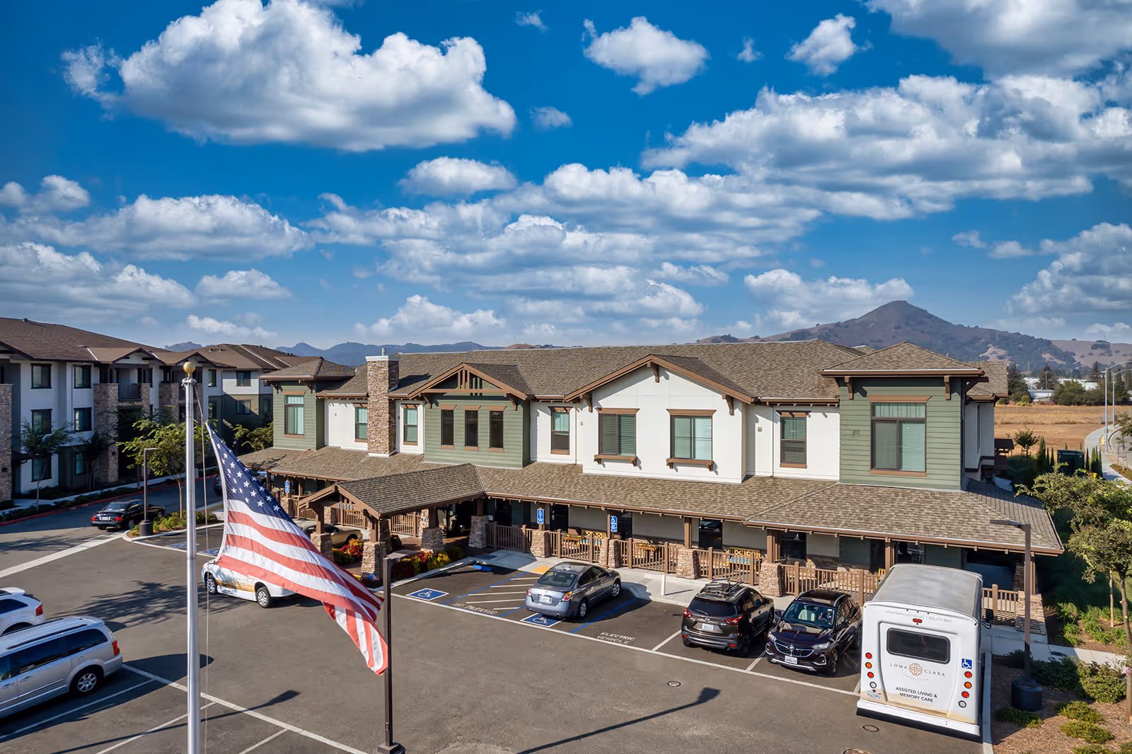 Two-story senior living building front with a parking lot, American flag, and distant hills under a partly cloudy sky.