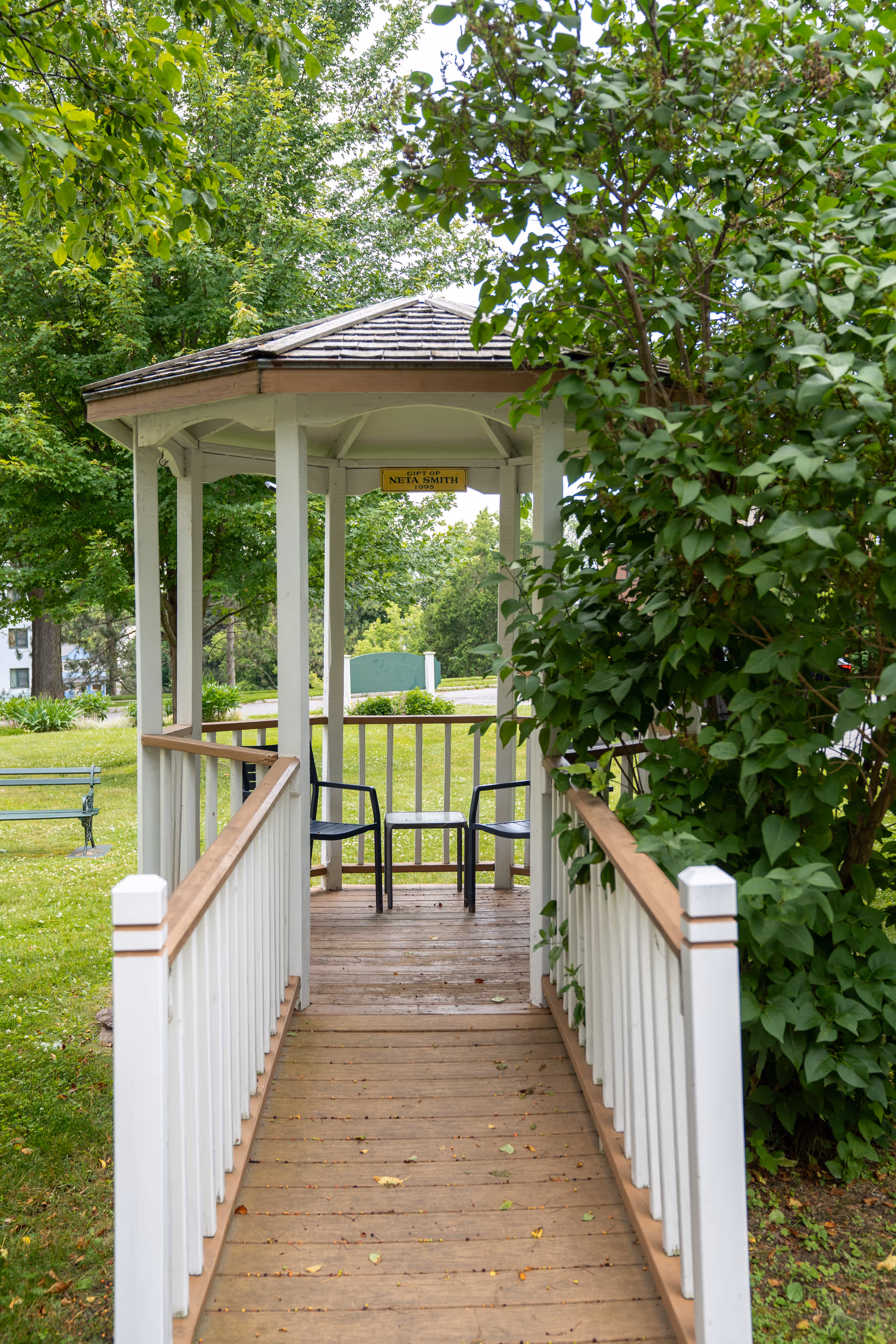 A wooden gazebo with a shingled roof and white railings surrounded by green trees and bushes. Two black chairs and a small table are inside the gazebo. A wooden ramp leads up to the gazebo. A small sign inside the gazebo reads 'Gift of Neta Smith'.