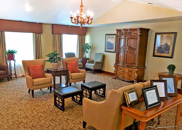 A cozy living room area with beige armchairs and small black stools arranged around a wooden side table. The room features a patterned carpet, a chandelier hanging from the ceiling, framed artwork on the walls, and a large wooden armoire. There are also several potted plants and a wooden table with framed documents or pictures on it.