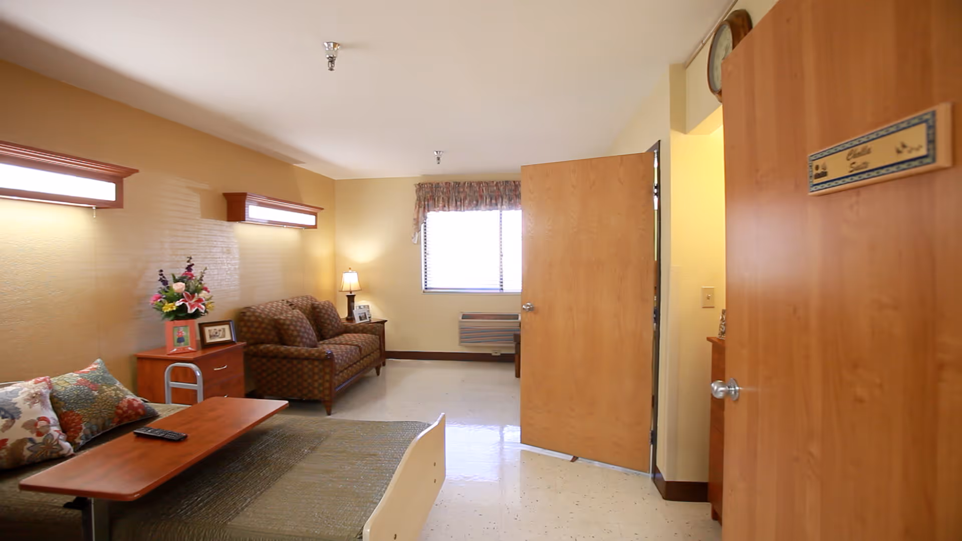 A bright and clean nursing center room with a hospital bed, a wooden overbed table, a patterned sofa, a small wooden side table with a lamp and framed photos, a window with blinds and floral valance, and an open wooden door with a sign that reads 'Cedar Suite'.