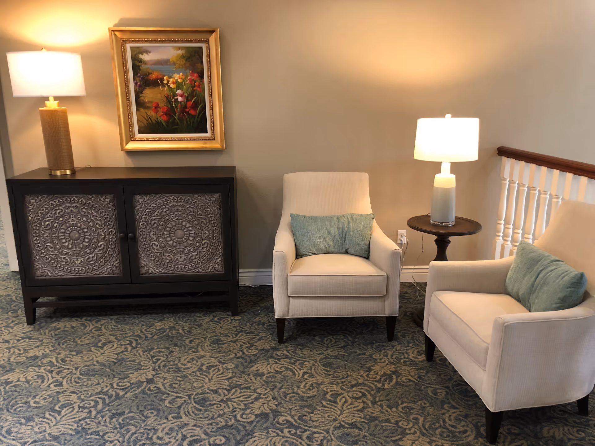 A cozy sitting area with two beige armchairs, each with a green cushion. Between the chairs is a small round wooden table with a white lamp. To the left is a dark wooden cabinet with ornate carved doors, topped with a lamp and a framed painting of a colorful garden scene on the wall above it. The floor is covered with a patterned carpet.