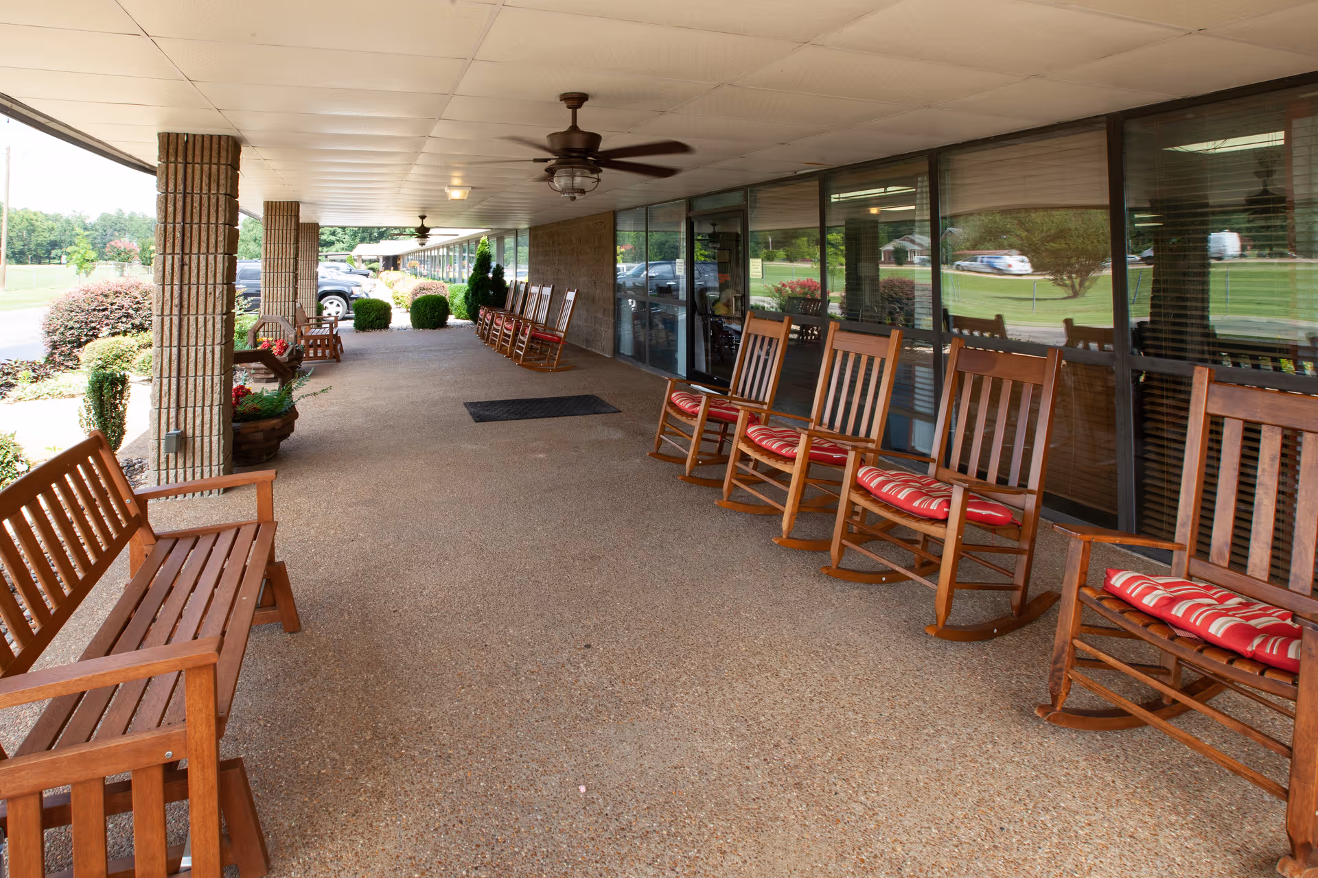 Covered outdoor patio area with wooden rocking chairs and benches lined up along the wall of a building. Ceiling fans are mounted on the white ceiling, and there are large windows reflecting the outdoor greenery and parked cars. The patio overlooks a landscaped area with bushes and trees.