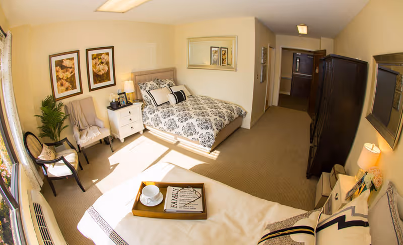 A bright and cozy bedroom in a senior living facility featuring a bed with patterned bedding, a nightstand with a lamp and photo frames, two chairs near a window with curtains, a potted plant, framed floral artwork on the wall, a large mirror, and a wooden dresser. Sunlight streams through the window, illuminating the room.