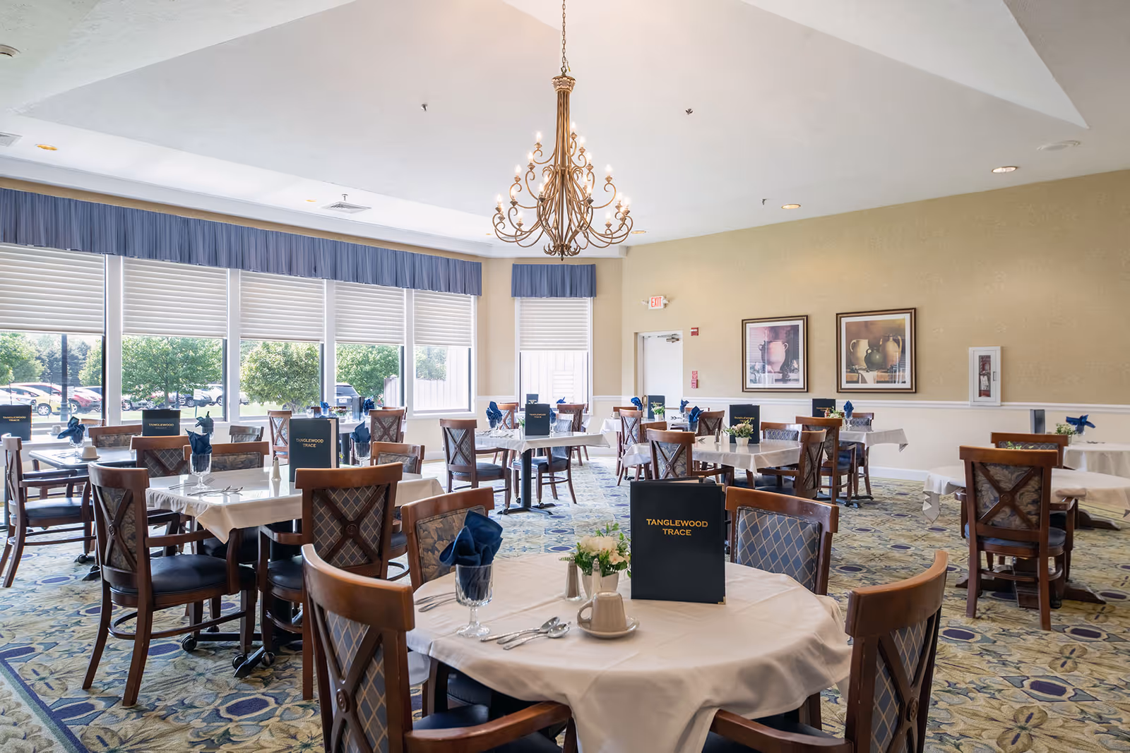 Spacious senior dining room with round tables set with white linens, chairs, menus labeled 'TANGLEWOOD TRACE', large windows, and a central chandelier.
