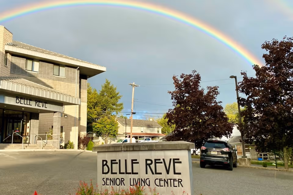 Front exterior of the Belle Rêve Senior Living Center with its entrance sign and a rainbow arching overhead.
