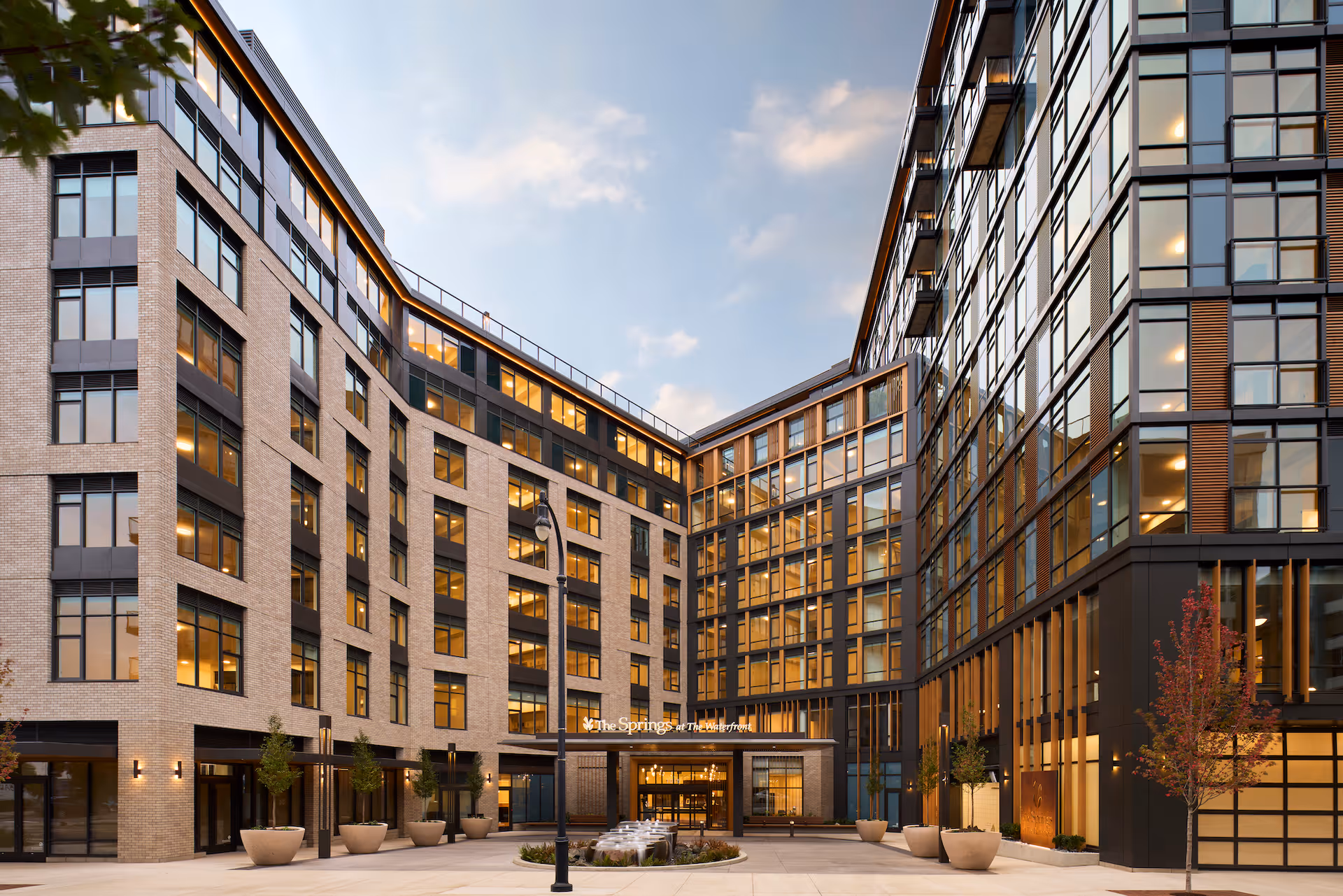 Exterior view of a modern multi-story senior living facility named The Springs at The Waterfront, featuring large windows, a central courtyard with planters and a water fountain, and warm lighting inside the building during dusk.