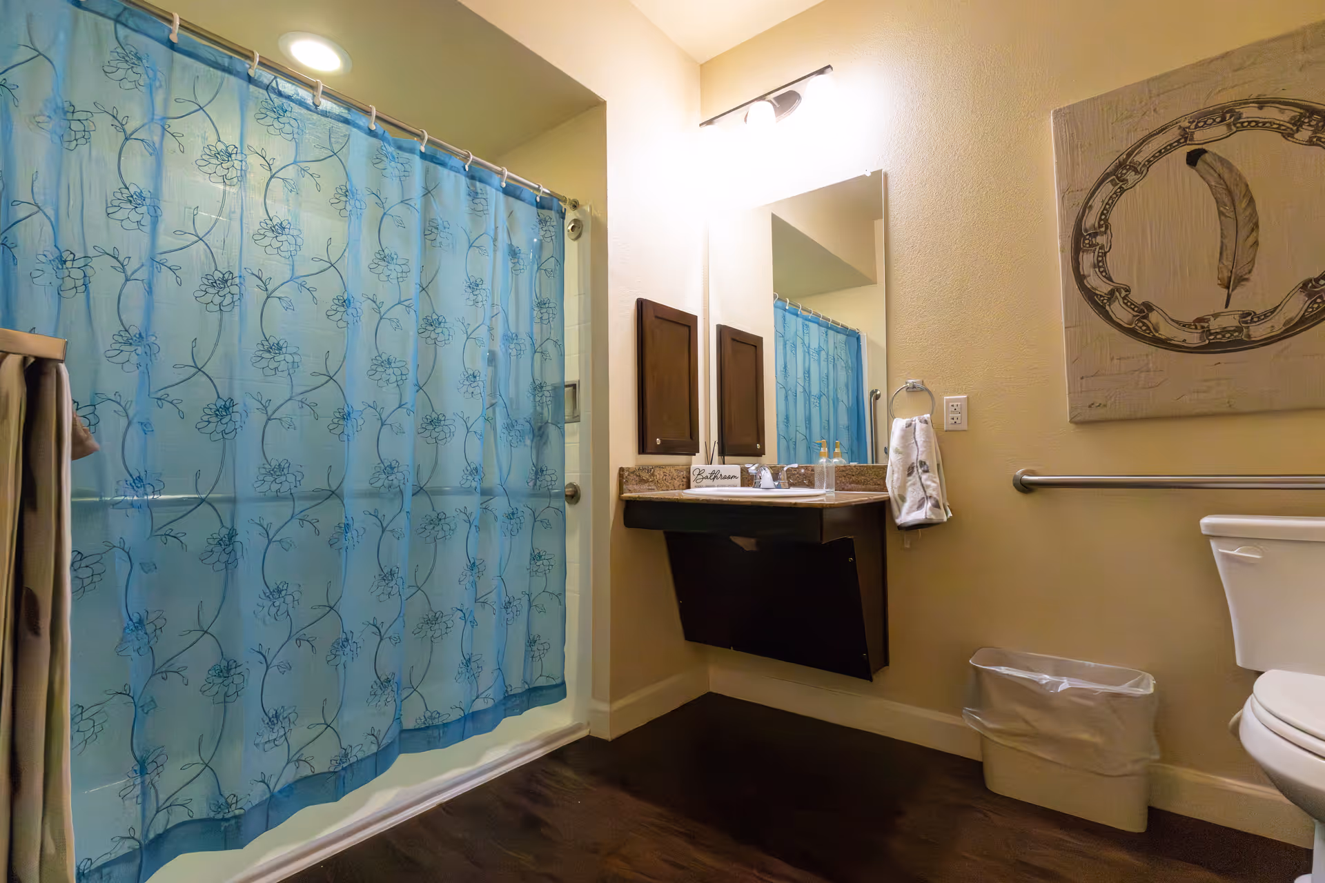 Bathroom with a blue floral shower curtain, a wall-mounted sink with a mirror above it, a towel hanging on a ring, a toilet, a trash bin, and a decorative wall art featuring a feather.