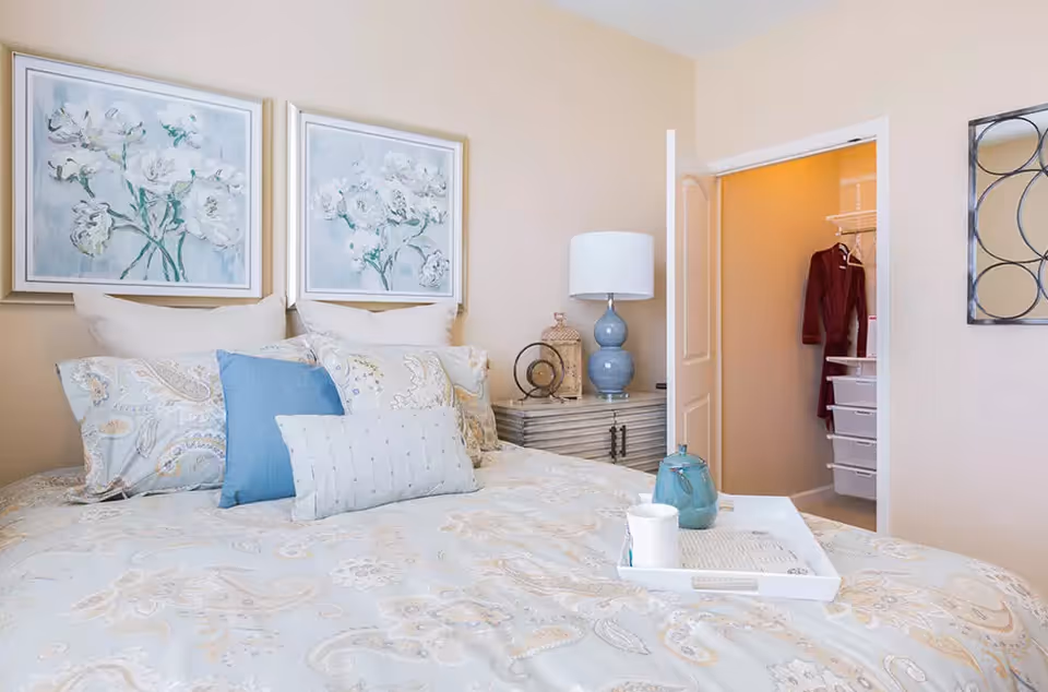 A cozy bedroom with a bed featuring patterned bedding and multiple pillows in shades of blue and beige. Above the bed are two framed floral paintings. To the right of the bed is a nightstand with a blue lamp and decorative items. An open door reveals a walk-in closet with a hanging maroon robe and white shelving units.