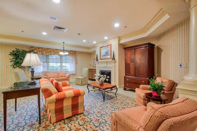A cozy senior living room with patterned carpet, striped and checkered upholstered armchairs and sofas, a wooden coffee table with a flower arrangement, a fireplace with a framed picture above it, a wooden armoire, and a window with floral valance curtains letting in natural light.