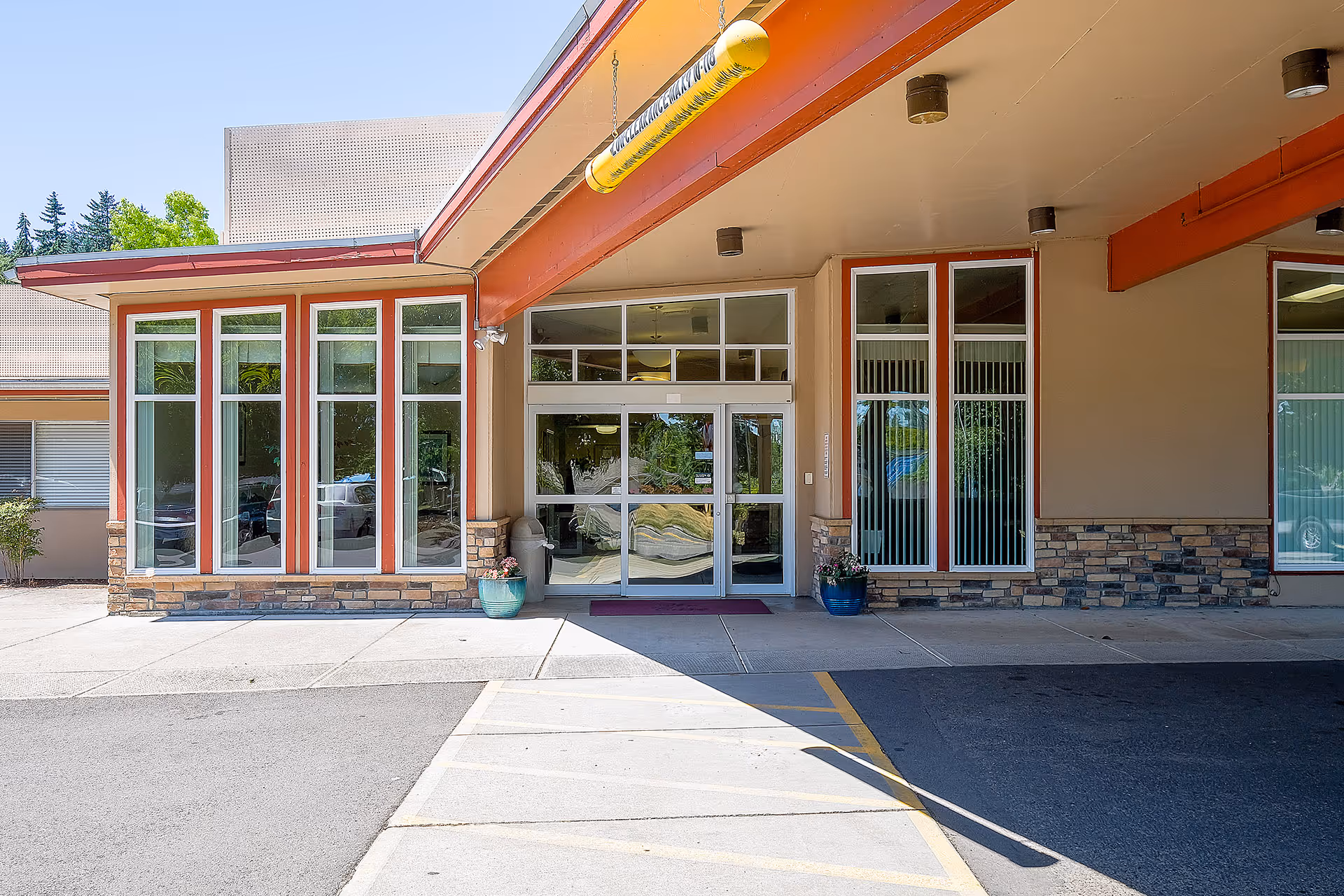 Entrance of Avamere Rehabilitation of Eugene showing glass double doors with large windows on either side, a covered walkway with an orange beam overhead, and potted plants near the entrance.