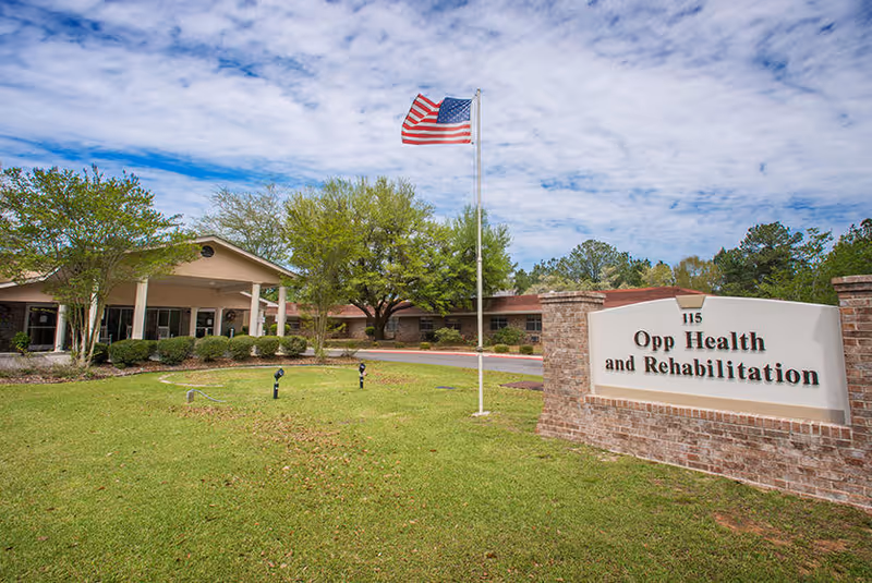 Exterior view of Opp Health and Rehabilitation facility with a brick sign displaying the name and address. A flagpole with an American flag stands on a well-maintained lawn in front of the building, which is surrounded by trees and shrubbery under a partly cloudy sky.