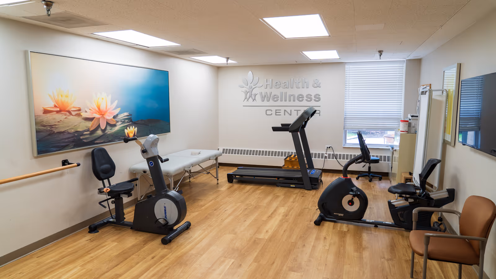 A health and wellness center room with exercise equipment including a recumbent bike, treadmill, and upright bike. The room has light wood flooring, white walls, a large window with blinds, a wall-mounted TV, a therapy table, and a large framed picture of water lilies on the wall.