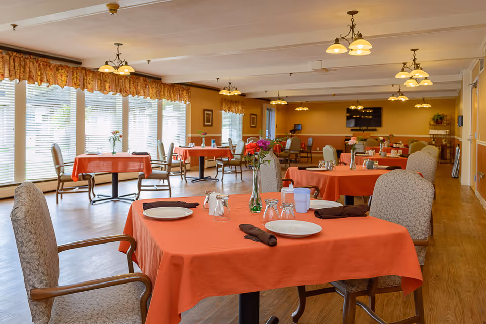 A dining room in a senior living facility with multiple tables covered in orange tablecloths. Each table is set with plates, glasses, napkins, and small flower vases. The room has large windows with blinds and floral valances, wooden flooring, and warm lighting from ceiling fixtures.