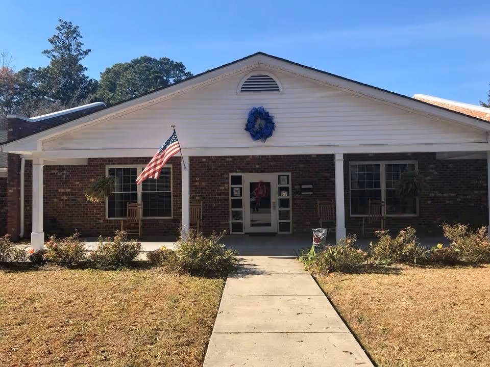 Front exterior view of a single-story brick building with a white triangular roof section. There is an American flag mounted on a white columned porch with rocking chairs on either side of the entrance. A blue wreath is displayed above the entrance door. The walkway leads up to the porch through a lawn with some bushes.