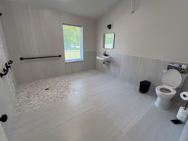 A spacious, accessible bathroom with a walk-in shower area featuring pebble-style flooring, a white toilet with the lid open, a small black trash bin, a wall-mounted sink with a mirror above it, and a window letting in natural light. The walls and floor are tiled in light neutral tones, and there are multiple black grab bars installed for support.