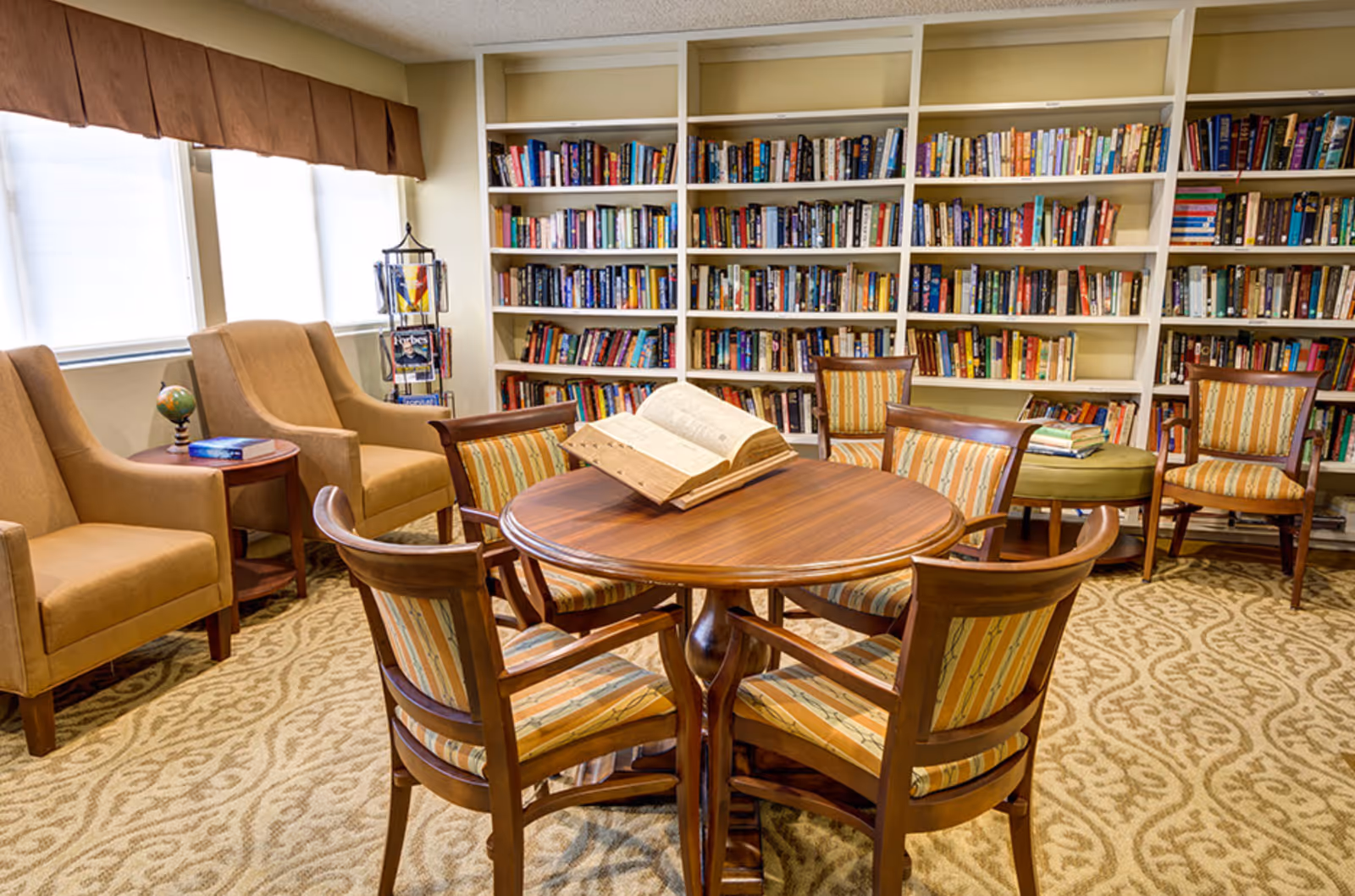 Cozy communal library room with a round wooden table and chairs, armchairs by the window, and bookshelves filled with books.