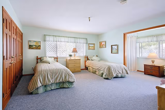 A spacious bedroom with two twin beds, each with striped bedding and a pillow. Between the beds is a wooden nightstand with a lamp and a small plant. The room has light blue walls, carpeted floor, wooden closet doors on the left, and several framed paintings on the walls. A window with sheer curtains lets in natural light.
