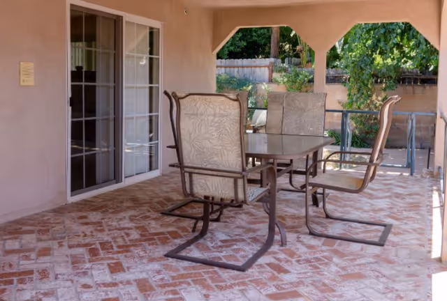 Outdoor patio with a glass table and four cushioned chairs beside a sliding door.
