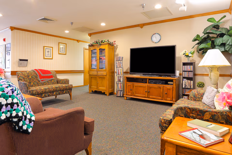 A cozy living room area in an assisted living facility featuring patterned armchairs and a loveseat with colorful crocheted blankets. There is a wooden TV stand with a large flat-screen television, a wooden cabinet with glass doors, a CD rack, and a tall plant in the corner. A table with books and a lamp is also visible.