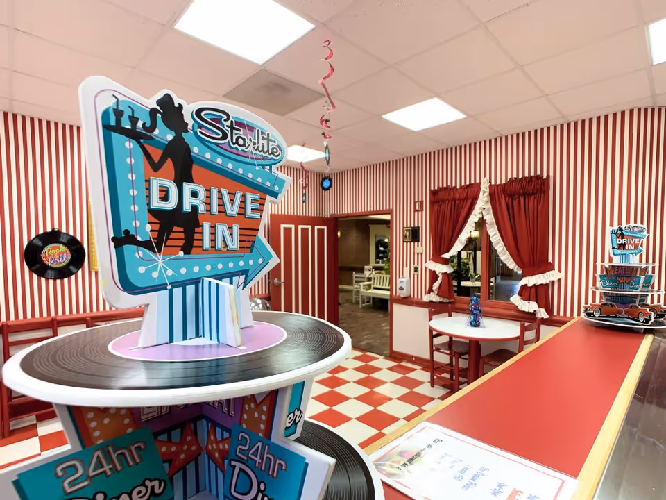 Interior of a retro-themed dining area with red and white striped walls and checkered floor tiles. A large decorative sign reading 'Starlite Drive In' is prominently displayed on a round table resembling a vinyl record. There is a red counter to the right with a small model car and another smaller Starlite Drive In sign. A round table with red chairs is positioned near a window with red curtains tied back with white ruffles. The ceiling has white tiles with fluorescent lights.