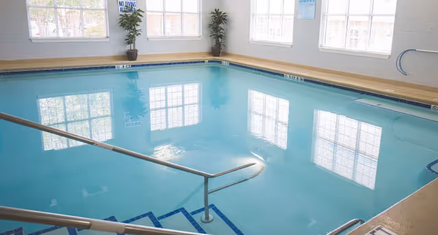 Indoor swimming pool with handrails and large windows reflecting on the water.
