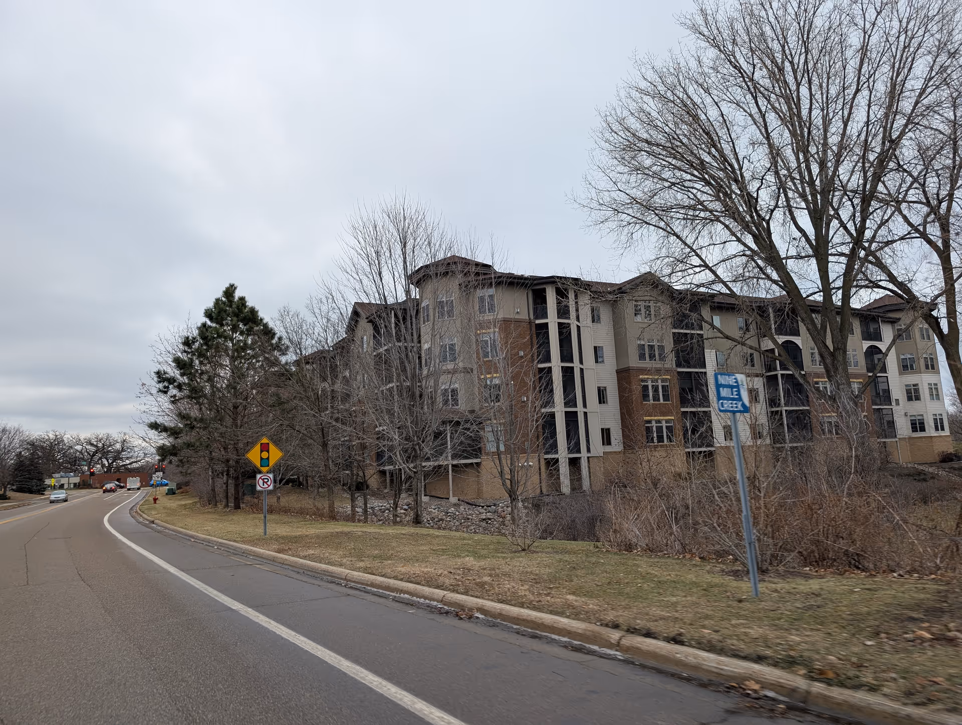 Exterior view of a multi-story residential building with beige and brown siding, surrounded by leafless trees and a grassy area next to a curved road under a cloudy sky.