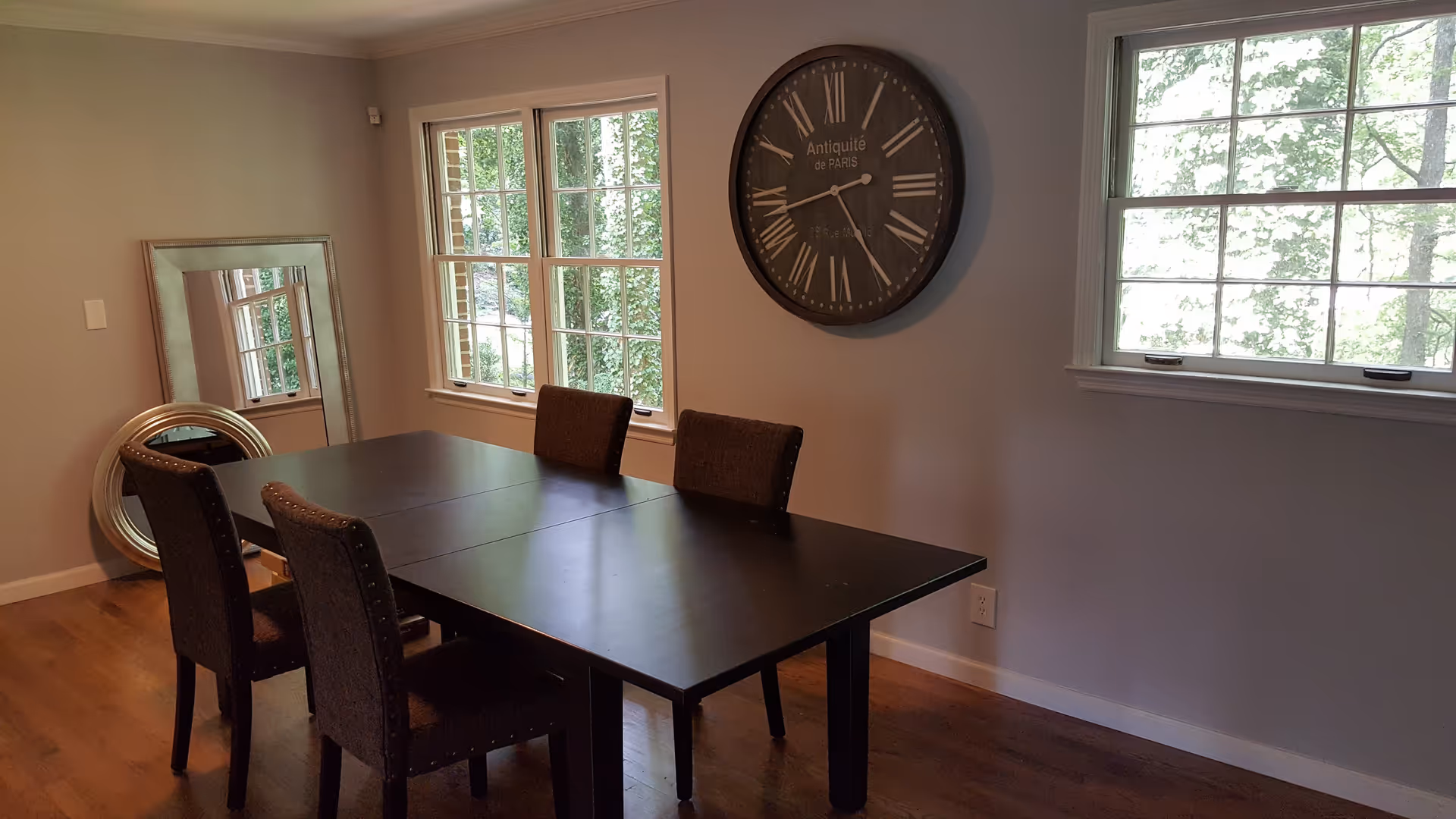 A dining room with a dark wooden table and four upholstered chairs. Two large windows let in natural light, and a large round wall clock with Roman numerals hangs on the wall. Two decorative mirrors, one rectangular and one round, lean against the wall in the corner. The floor is wooden.