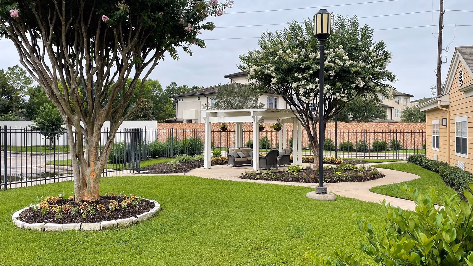 Outdoor garden area at Atria Westchase featuring a green lawn, a large tree with a circular stone border, a white pergola with seating underneath, a lamp post, and surrounding buildings and fencing.