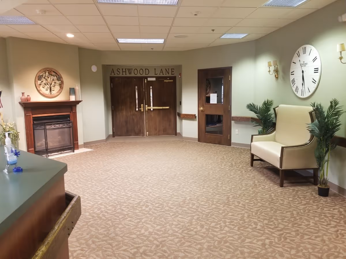 A hallway area in a senior living facility with double wooden doors labeled 'ASHWOOD LANE'. The space features a beige patterned carpet, a wooden fireplace mantel with decorative items, a large wall clock, a cream-colored bench with wooden arms, two potted plants, and wall sconces. The ceiling has recessed lighting and a drop ceiling with fluorescent lights.