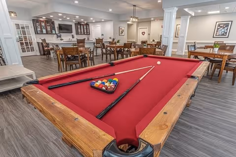 A spacious recreational room featuring a red felt pool table with cues and balls arranged for a game. In the background, there are multiple wooden tables and chairs arranged for dining or socializing, a cabinet with glass doors, a television mounted on the wall, and framed artwork. The room has wood flooring and neutral-colored walls with white trim and columns.