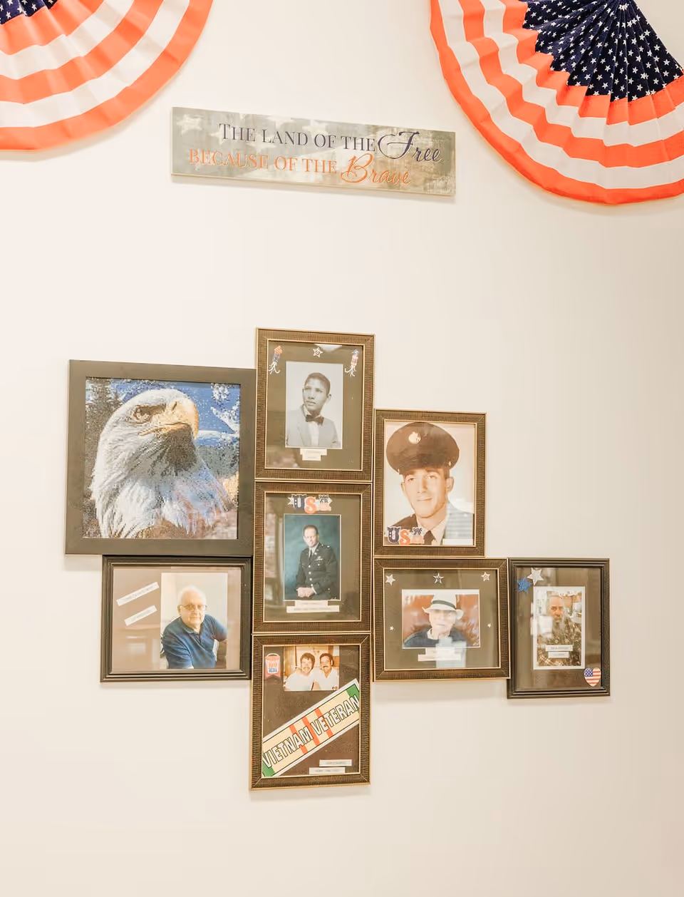 A wall display featuring framed photographs of veterans, including portraits of men in military uniforms and casual attire, a framed image of a bald eagle, and a sign above that reads 'THE LAND OF THE Free BECAUSE OF THE Brave'. Two patriotic red, white, and blue fan decorations are partially visible at the top corners of the image.