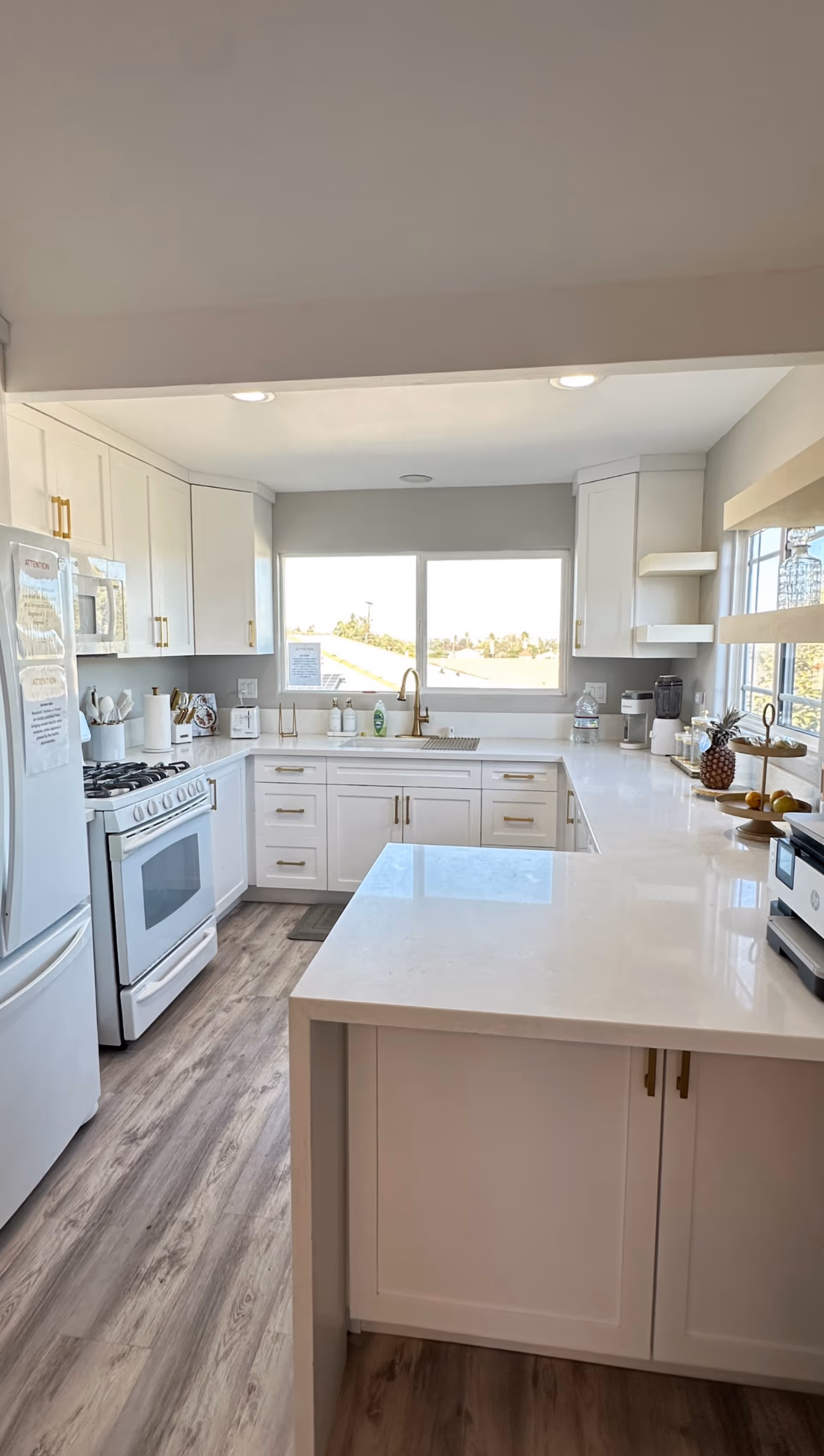 Bright and modern kitchen with white cabinetry, a white countertop island, a gas stove, a refrigerator, and a large window above the sink letting in natural light. The kitchen has wood-style flooring and various kitchen appliances and utensils on the counters.