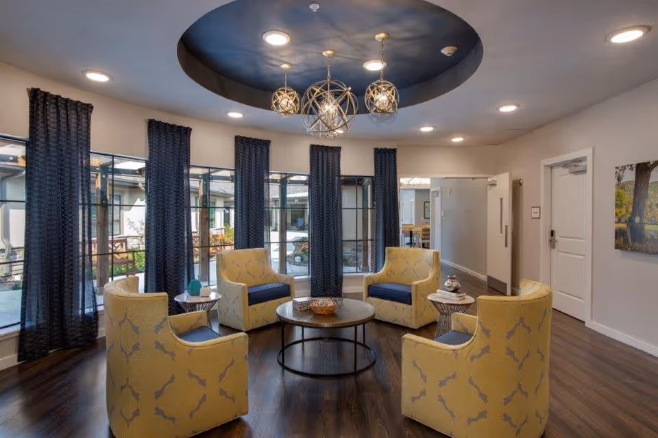A cozy seating area with five yellow armchairs arranged in a circle around a round wooden coffee table. The room has large windows with dark curtains, allowing natural light to enter. The ceiling features a recessed circular design with a modern chandelier hanging in the center. The floor is wooden, and there are two small side tables with decorative items. A hallway and a door are visible in the background.