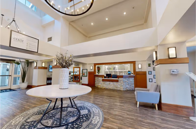 Spacious and well-lit reception area of Cogir of Vacaville featuring a round table with a vase of flowers in the center on a patterned rug, a modern stone and wood reception desk with two chairs behind it, light wood flooring, high ceilings with large windows, and comfortable seating to the right.