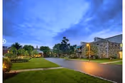 Evening view of a paved driveway leading to a stone building with outdoor lighting and landscaped greenery under a cloudy sky.