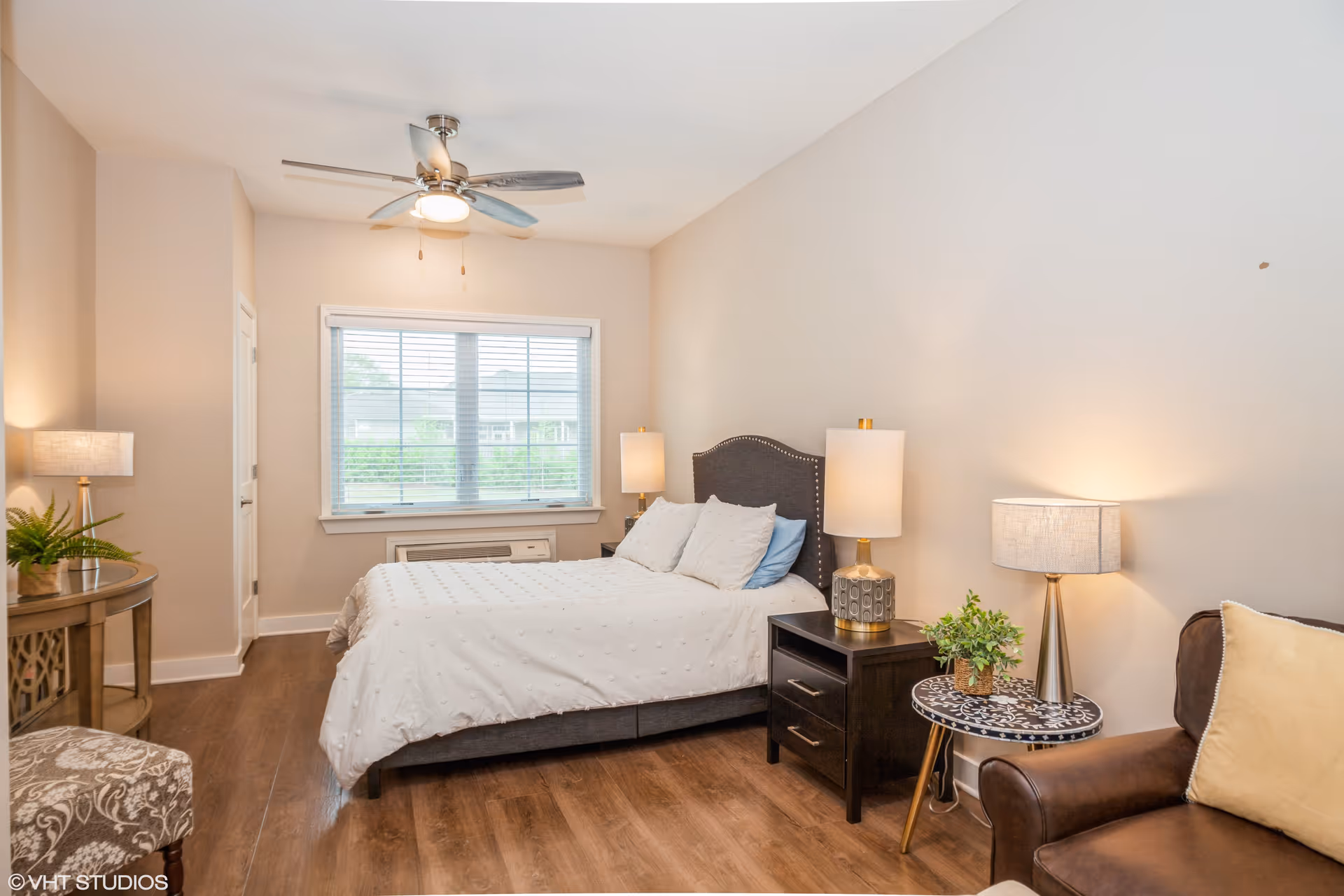 A bright and cozy bedroom with a large window covered by white blinds. The room features a bed with a white textured comforter and multiple pillows, two bedside tables with lamps, a ceiling fan with light, a small round table with a plant and lamp, a brown leather armchair with a beige pillow, and a decorative chair with a patterned fabric. The floor is wooden, and the walls are painted a light beige color.