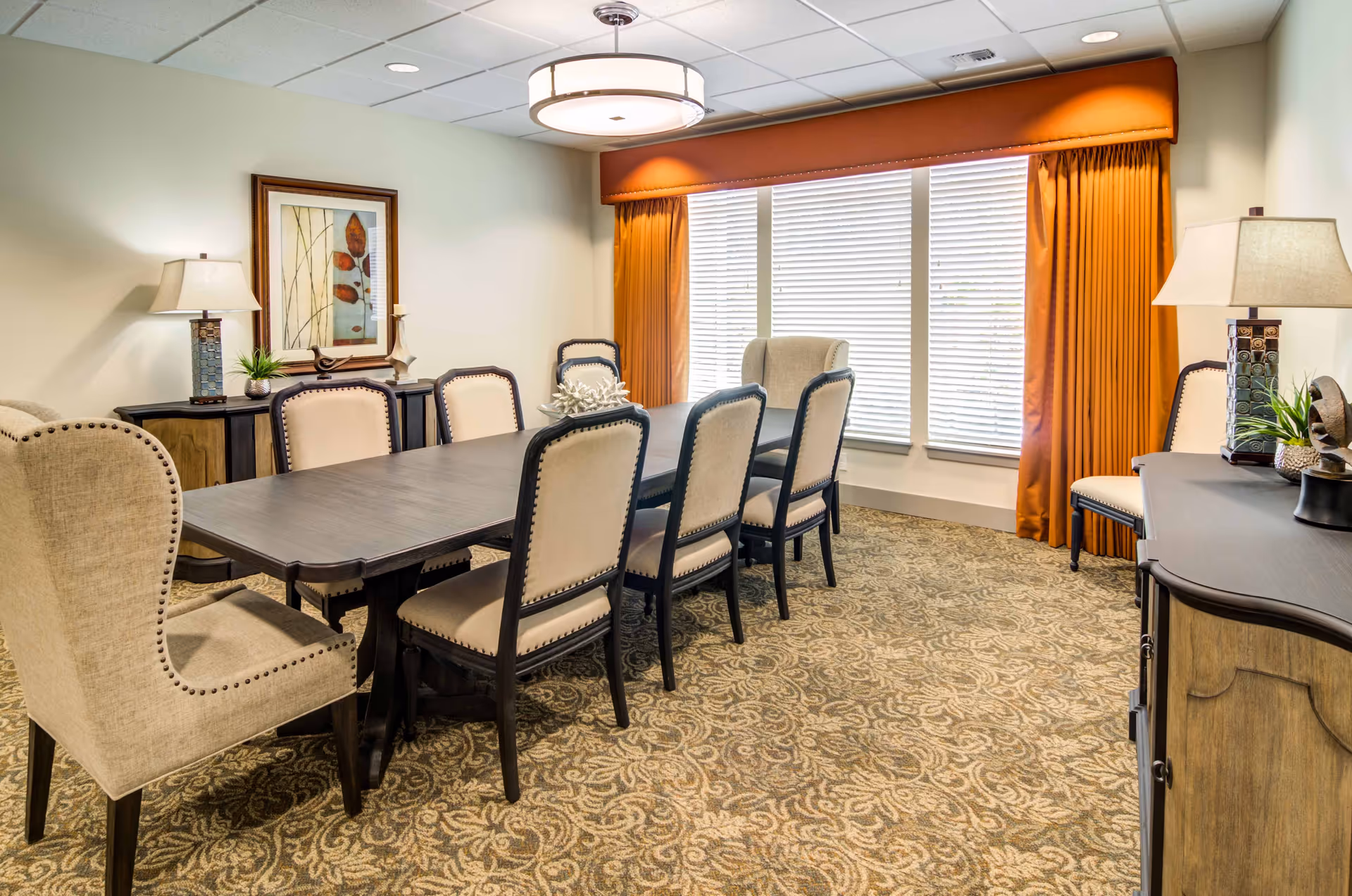 A well-lit dining room with a long dark wooden table surrounded by eight upholstered chairs. The room features patterned carpet, two table lamps on sideboards, a framed artwork on the wall, and large windows with orange curtains and white blinds.