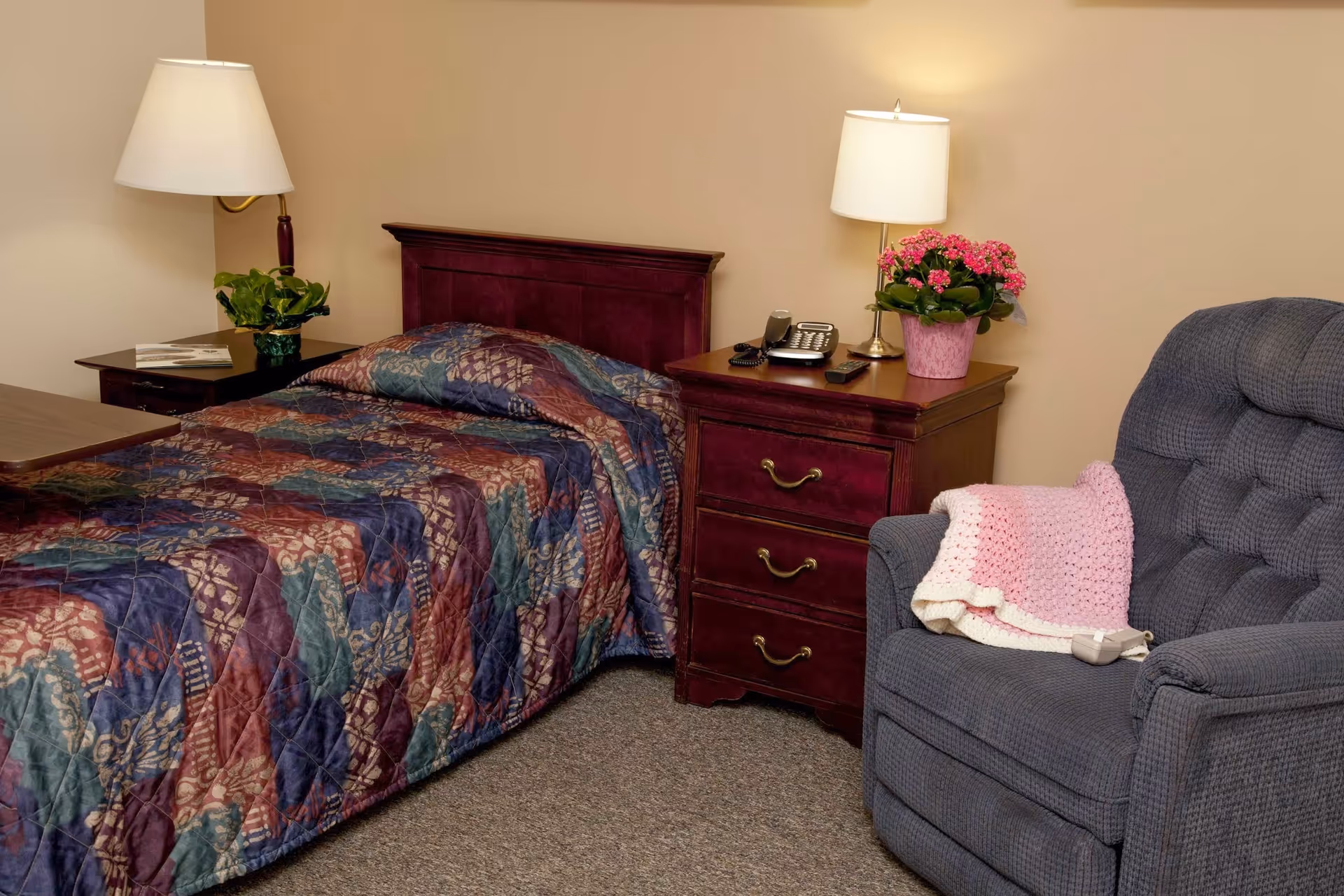 A small bedroom with a single bed covered by a patterned quilt, a wooden nightstand with a lamp, phone and potted plant, and a blue upholstered recliner with a pink blanket.