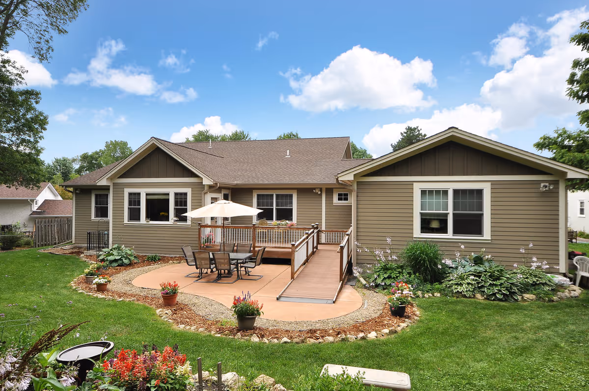 Back view of a single-story house with beige siding and a brown roof, featuring a patio area with a table, chairs, and an umbrella. The patio is surrounded by a small garden with various plants and flowers, and a wooden ramp leads from the patio to the backyard lawn under a partly cloudy blue sky.