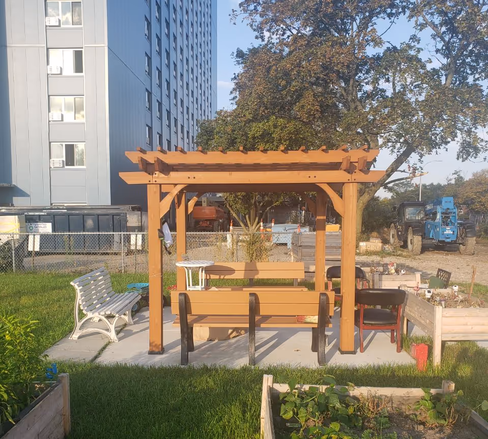 A wooden pergola with benches and chairs in a small garden with raised planters and an apartment building and construction equipment in the background.