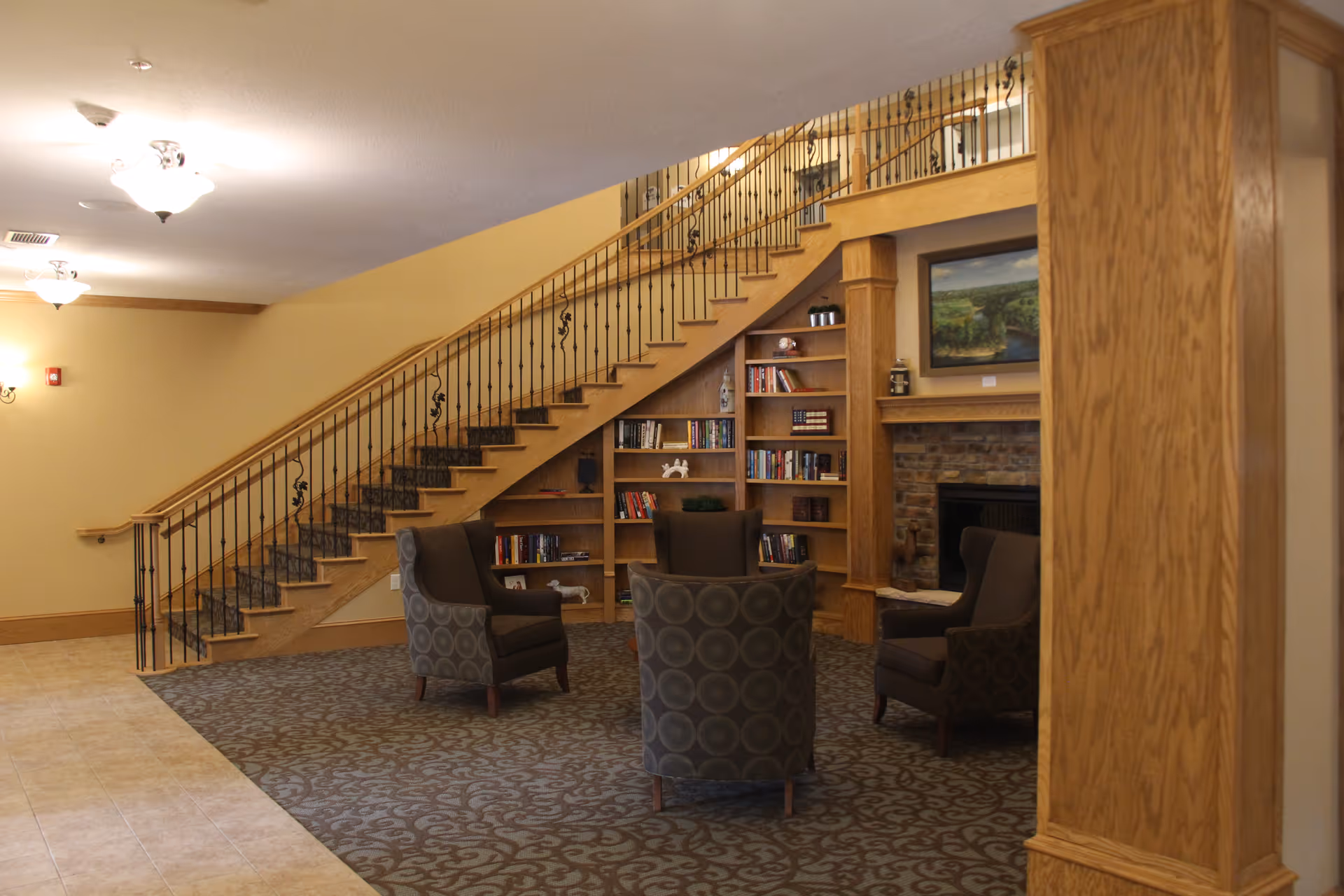 Interior view of a senior living facility lounge area featuring a carpeted seating space with four patterned armchairs arranged around a fireplace. Behind the chairs is a wooden bookshelf filled with books and decorative items. A staircase with a wooden handrail and black iron balusters ascends along the wall. The walls are painted beige and the floor near the seating area is carpeted with a patterned design, while the adjacent floor is tiled.