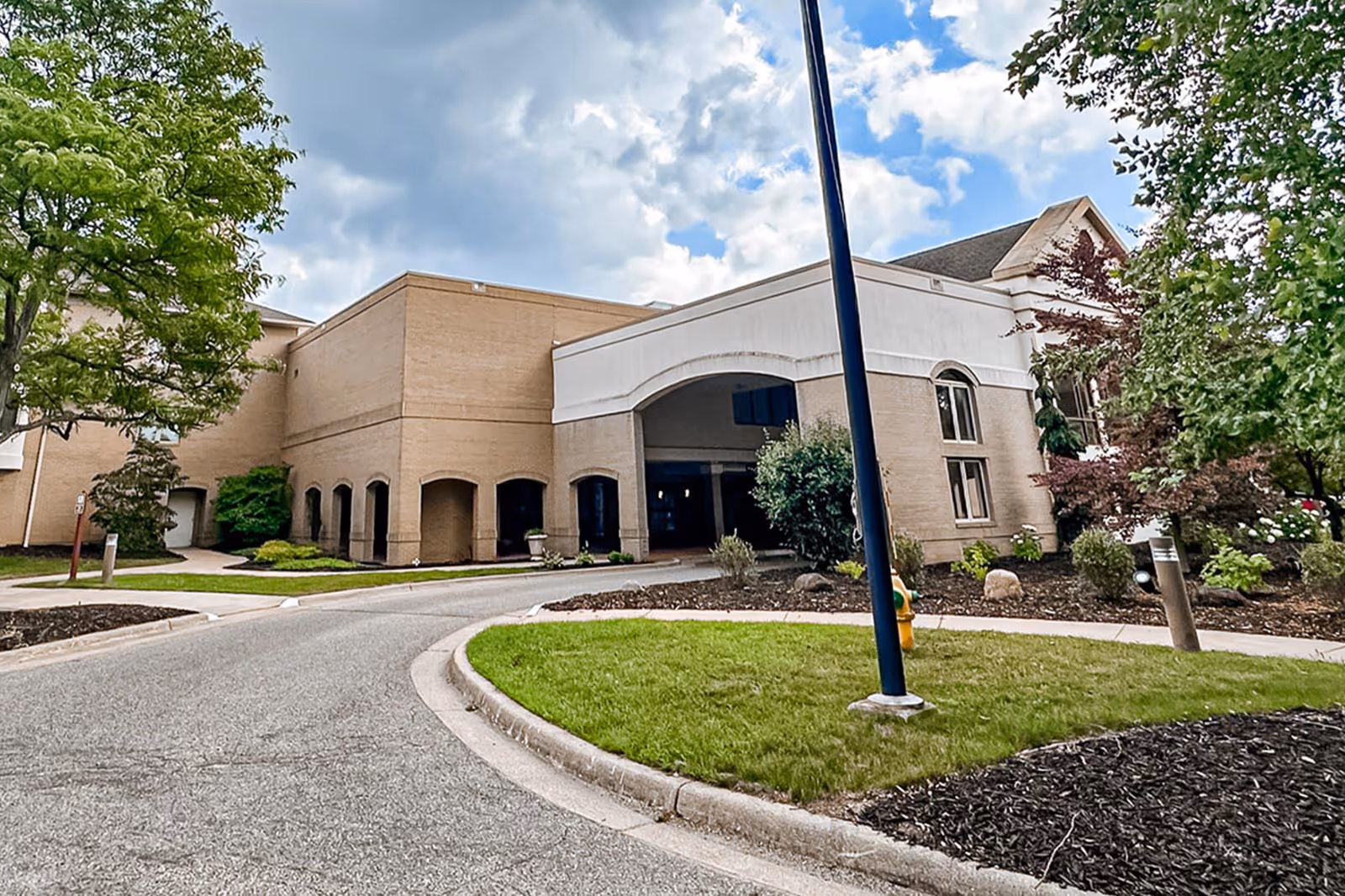Front entrance and drive of a beige brick senior living building with landscaped grounds and a cloudy sky.