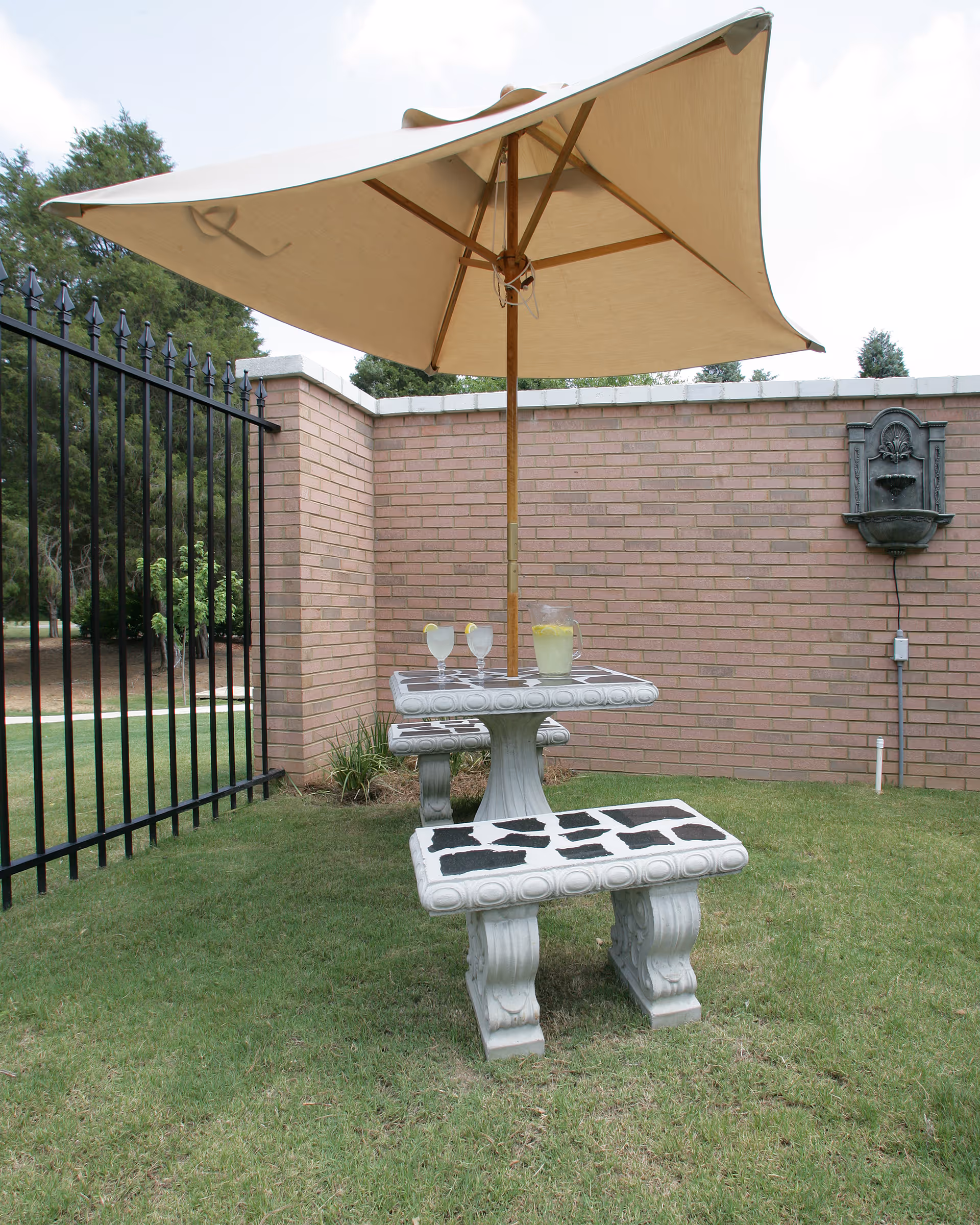 Outdoor seating area with a stone table and two benches on grass, shaded by a large beige umbrella. On the table are two glasses of lemonade with lemon slices and a pitcher of lemonade. The area is enclosed by a brick wall and a black metal fence, with trees visible in the background.