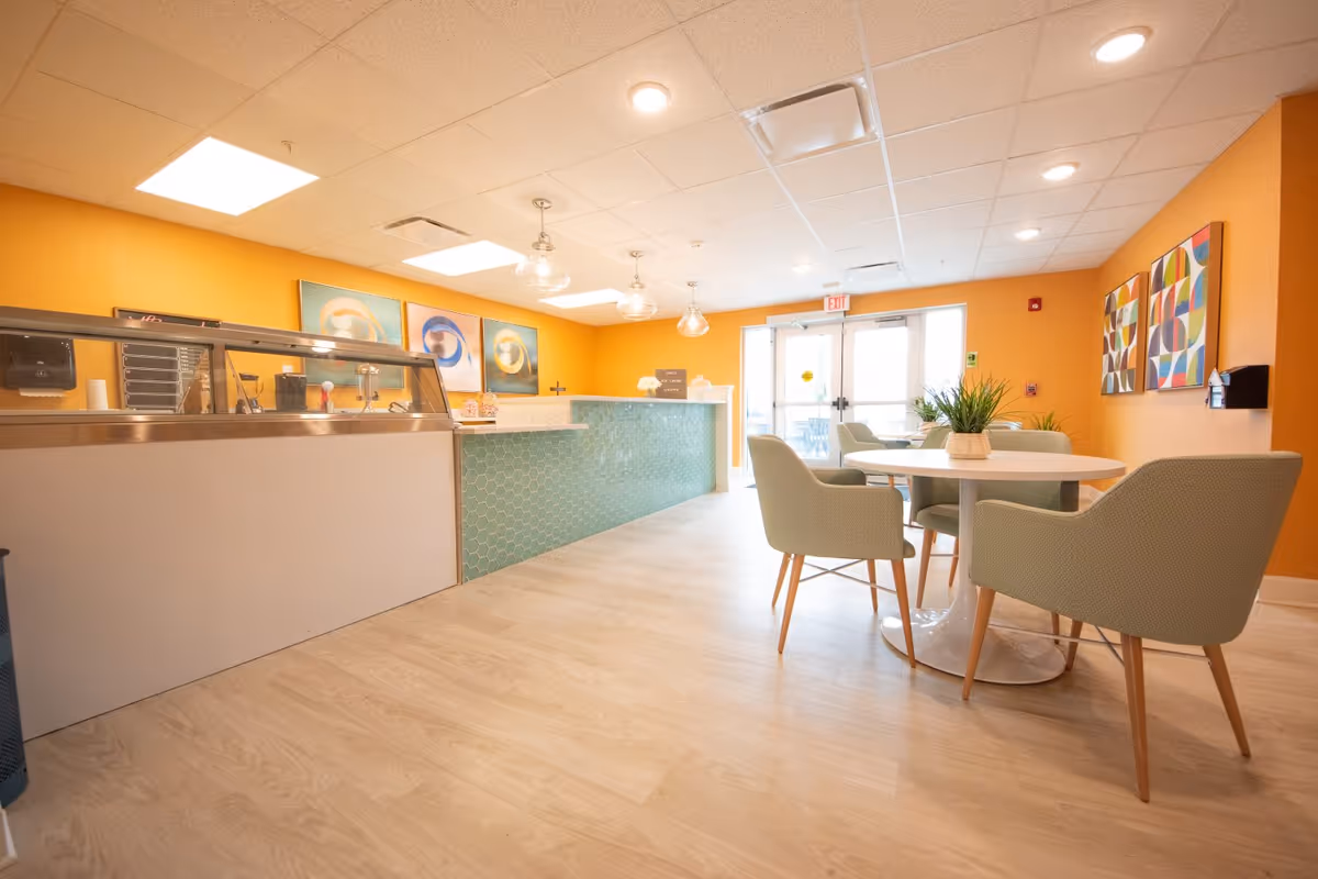 Bright common dining and reception area with a round table and chairs, a green-tiled service counter, and orange walls with artwork.