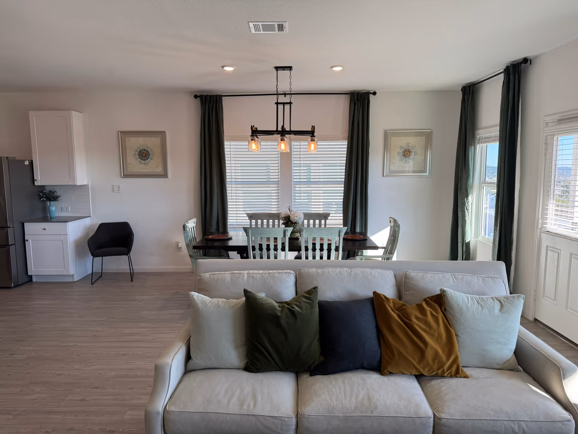 A bright and modern living and dining area with a beige sofa adorned with four cushions in white, green, navy, and mustard colors. Behind the sofa is a dark wooden dining table with six white chairs, a flower centerpiece, and a hanging light fixture with three bulbs. The room has light wood flooring, white walls, two framed artworks, large windows with green curtains, and a partial view of a kitchen area with white cabinets and a stainless steel refrigerator.