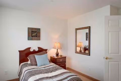 A small bedroom with a single bed featuring a wooden headboard, striped bedding, and three pillows. Next to the bed is a wooden nightstand with a lamp, a phone, and a small decorative item. A framed mirror hangs on the wall above the nightstand, reflecting the lamp. The walls are painted white, and there is a piece of artwork hanging above the bed. The door to the room is partially open.