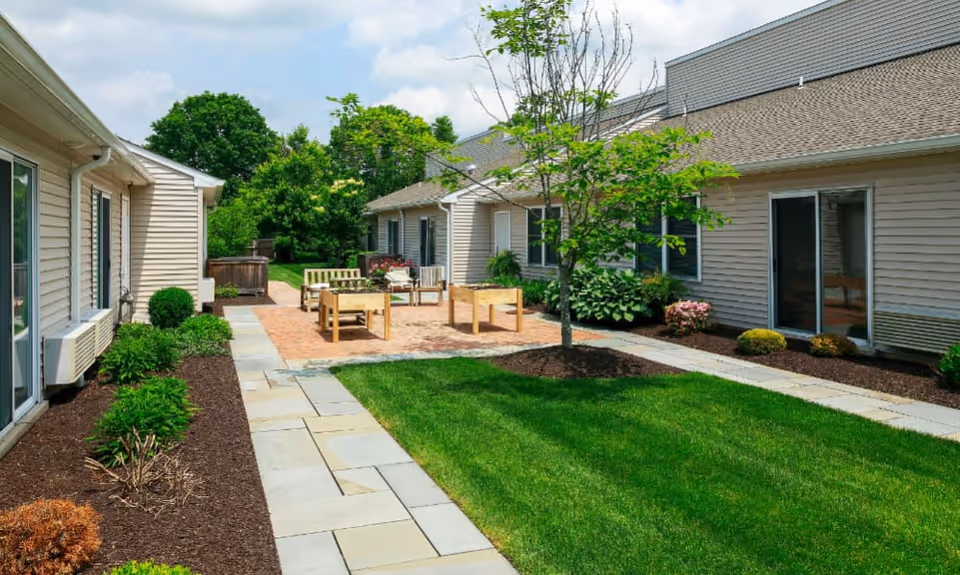 Outdoor courtyard area at Traditions of Lansdale featuring a paved walkway, green lawn, small trees, shrubs, and patio furniture including chairs and tables between beige residential buildings under a partly cloudy sky.