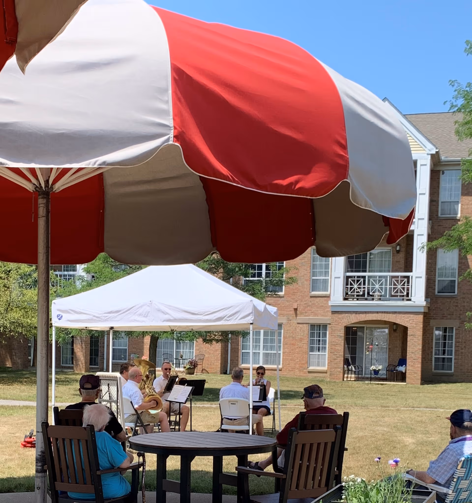 Outdoor scene at The Parkvue Community showing elderly people seated on chairs under a large red and white umbrella, watching a small brass band performing under a white canopy tent on a sunny day with a brick building in the background.
