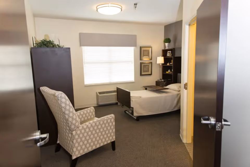 A private senior living bedroom with a single adjustable bed, patterned armchair, shelving, and a window with blinds.